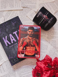 Load image into Gallery viewer, A red Tarot Card titled "The Archer" with the image of a shirtless man on it, on top of a book titled Kate by Tate James with flowers, book pages, and a candle in the background.