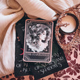 Load image into Gallery viewer, A Tarot Card "The Starblessed" with an image of a celestial woman printed on it on top of a book stack from the series A Kingdom of Stars and Shadows by Holly Renee with flowers and other books in the background.