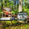 Load image into Gallery viewer, A postage stamp featuring artwork of a romantic scene, on top of a book titled The Simple Wild by K.A. Tucker, on top of a mossy log with a forest in the background. A wooden sign with a mountain and forest printed on it with the words "The Simple Wild' in green on it.