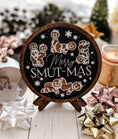 Load image into Gallery viewer, Round, wooden sign with a navy blue background and dark woodgrain frame. Featuring text that reads "Merry Smut-mas" surrounded by gingerbread men and women in smutty positions. ©FireDrake Artistry 2024.