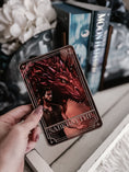 Load image into Gallery viewer, A 'Sabersythe' Tarot Card featuring an illustration of a man and red dragon being held in a hand with books and flowers on a table in the background.