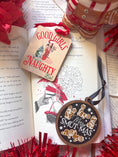 Load image into Gallery viewer, A rectangular wooden ornament featuring a Christmas tree of books next to a girl in a Santa hat tied up with Christmas lights surrounded by text that reads 'Good Girls Naughty List'. © FireDrake Artistry 2023