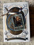 Load image into Gallery viewer, Tarot Card ornament titled "The Shield Maiden" with silver ribbon and book underneath.
