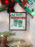 Load image into Gallery viewer, Decorative sign with a dark wood frame, 'Plot Twists & Peppermint' text, with skeleton hands holding hot chocolate, on a white background. © FireDrake Artistry 2025
