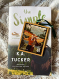 Load image into Gallery viewer, Stamp ornament with the words "Alaska Wild" and a couple in front of a plane on it. Book titled "The Simple Wild" in the background.