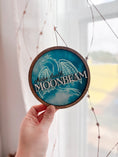 Load image into Gallery viewer, Round wooden sign with 'MOONBEAM' text and dragon design held in hand.