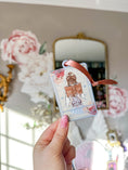 Load image into Gallery viewer, "The Reader" Tarot Ornament, Option 1. A white ornament with a lavender border featuring a girl reading a book surrounded by flowers. Text reads "The Reader." © FireDrake Artistry® 2024.