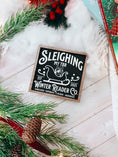 Load image into Gallery viewer, Wooden shelf sign with a black background and woodgrain frame. A white sleigh with presents, surrounded by white text reading 'Sleighing My TBR' and "Winter Reader Co. EST 2025." © FireDrake Artistry 2025