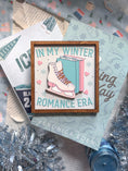 Load image into Gallery viewer, A wooden sign with a white background and woodgrain frame displaying the phrase 'IN MY WINTER ROMANCE ERA' surrounded by winter-themed illustrations such as a ice skate, snowflakes, and hearts. © FireDrake Artistry 2024