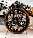 Load image into Gallery viewer, Round, wooden ornament with a navy blue background and dark woodgrain frame. Featuring text that reads "Merry Smut-mas" surrounded by gingerbread men and women in smutty positions. ©FireDrake Artistry 2024.