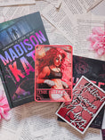Load image into Gallery viewer, A pink tarot card labeled "The Hellcat" on top of a book titled Madison Kate by Tate James with a shelf sign, books, flowers, and book pages in the background.