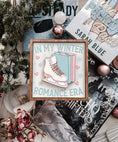 Load image into Gallery viewer, A wooden sign with a white background and woodgrain frame displaying the phrase 'IN MY WINTER ROMANCE ERA' surrounded by winter-themed illustrations such as a ice skate, snowflakes, and hearts. © FireDrake Artistry 2024