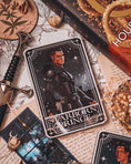 Load image into Gallery viewer, Tarot Card titled "Starboard Prince" with an image of a warrior with a sword printed on it, in front of books by Sarah J. Mass, flowers, and other bookish trinkets.