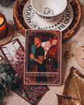 Load image into Gallery viewer, Tarot Card titled "Shadowbeast" with an image of a romantic couple printed on it, with book themed trinkets in the background.