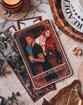 Load image into Gallery viewer, Tarot Card titled "Shadowbeast" with an image of a romantic couple printed on it, with book themed trinkets in the background.