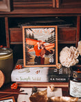 Load image into Gallery viewer, A postage stamp featuring artwork of a romantic scene, on top of a book titled The Simple Wild by K.A. Tucker, on a vintage desk with flowers and candles in the background.