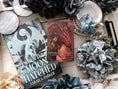 Load image into Gallery viewer, A 'Sabersythe' Tarot Card featuring an illustration of a red dragon, placed on a table with various decorative items including a white flower and other small trinkets.