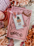 Load image into Gallery viewer, "The Reader" Tarot Ornament, Option 3. A white ornament with a lavender border featuring a girl reading a book surrounded by flowers. Text reads "The Reader." © FireDrake Artistry® 2024.