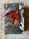 Load image into Gallery viewer, Tarot Card ornament titled "The Sabersythe" with maroon ribbon and book underneath.