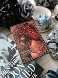 Load image into Gallery viewer, A 'Sabersythe' Tarot Card featuring an illustration of a red dragon, placed on a table with various decorative items including a white flower and other small trinkets.