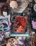 Load image into Gallery viewer, A 'Sabersythe' Tarot Card featuring an illustration of a red dragon, placed on a table with various decorative items including a white flower and other small trinkets.