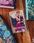 Load image into Gallery viewer, Tarot Card titled "The Lovers" with an image of a fantasy couple in a romantic embrace, on a wood floor surrounded by colorful books