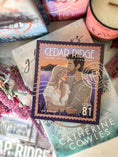 Load image into Gallery viewer, A postage stamp featuring artwork of a romantic scene, on top of a stack of books with a book stack and candle in the background.