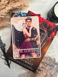 Load image into Gallery viewer, Tarot Card titled "The Lovers" with an image of a fantasy couple in a romantic embrace, on top of a stack of books with bookish trinkets in the background.