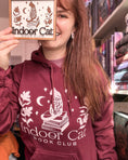 Load image into Gallery viewer, Woman next to bookshelf wearing indoor cat hoodie in maroon with white design