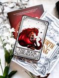 Load image into Gallery viewer, Tarot Card titled "The Book Dragon" with an image of a girl reading a book with a red dragon, , on top of a stack of books with flowers in the background.