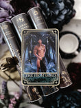 Load image into Gallery viewer, Tarot Card titled "The Highlord" with a single, shirtless man printed on the card, on top of a stack of books with flowers and bookish trinkets in the background.