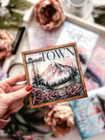 Load image into Gallery viewer, Sign with "Small Town Romance" and a mountain with trees and a lake printed on it. Books and flowers in the background.