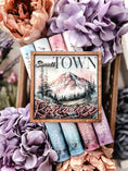 Load image into Gallery viewer, Sign with "Small Town Romance" and a mountain with trees and a lake printed on it. Books and flowers in the background.