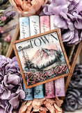 Load image into Gallery viewer, Sign with "Small Town Romance" and a mountain with trees and a lake printed on it. Books and flowers in the background.