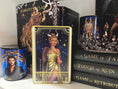 Load image into Gallery viewer, Tarot Card titled "The Goddess " with an image of a woman with horns in a gold dress, on a bookshelf with books in the background.