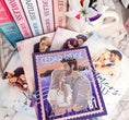 Load image into Gallery viewer, A postage stamp featuring artwork of a romantic scene, on top of a stack of books with a book stack and mug with flowers in the background.
