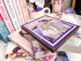 Load image into Gallery viewer, A postage stamp featuring artwork of a romantic scene, on top of a stack of books with a book stack and mug with flowers in the background.