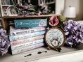 Load image into Gallery viewer, Round wooden sign with the words "Lost & Found" and a compass with flowers printed on it. Sign is placed on a wooden easel. Flowers and books in the background