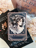 Load image into Gallery viewer, A Tarot Card "The Starblessed" with an image of a celestial woman printed on it on top of a book stack from the series A Kingdom of Stars and Shadows by Holly Renee with flowers and other books in the background.