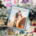 Load image into Gallery viewer, A postage stamp featuring artwork of a romantic scene, with a books and decorative flowers in the background.