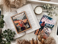 Load image into Gallery viewer, A postage stamp featuring artwork of a romantic scene, on top of an open book with another book titled The Simple Wild by K.A. Tucker, on top of a white wooden tray with a candle and greenery on it.