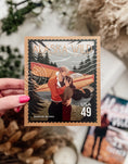 Load image into Gallery viewer, A hand holding a postage stamp featuring artwork of a romantic scene with flowers and books in the background