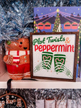 Load image into Gallery viewer, Decorative sign with a dark wood frame, 'Plot Twists & Peppermint' text, with skeleton hands holding hot chocolate, on a white background. © FireDrake Artistry 2025