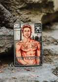 Load image into Gallery viewer, A grey tarot card labeled "The Trainer" with a shirtless man printed on it, on a stone step