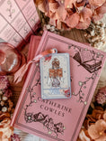 Load image into Gallery viewer, "The Reader" Tarot Ornament, Option 1. A white ornament with a lavender border featuring a girl reading a book surrounded by flowers. Text reads "The Reader." © FireDrake Artistry® 2024.