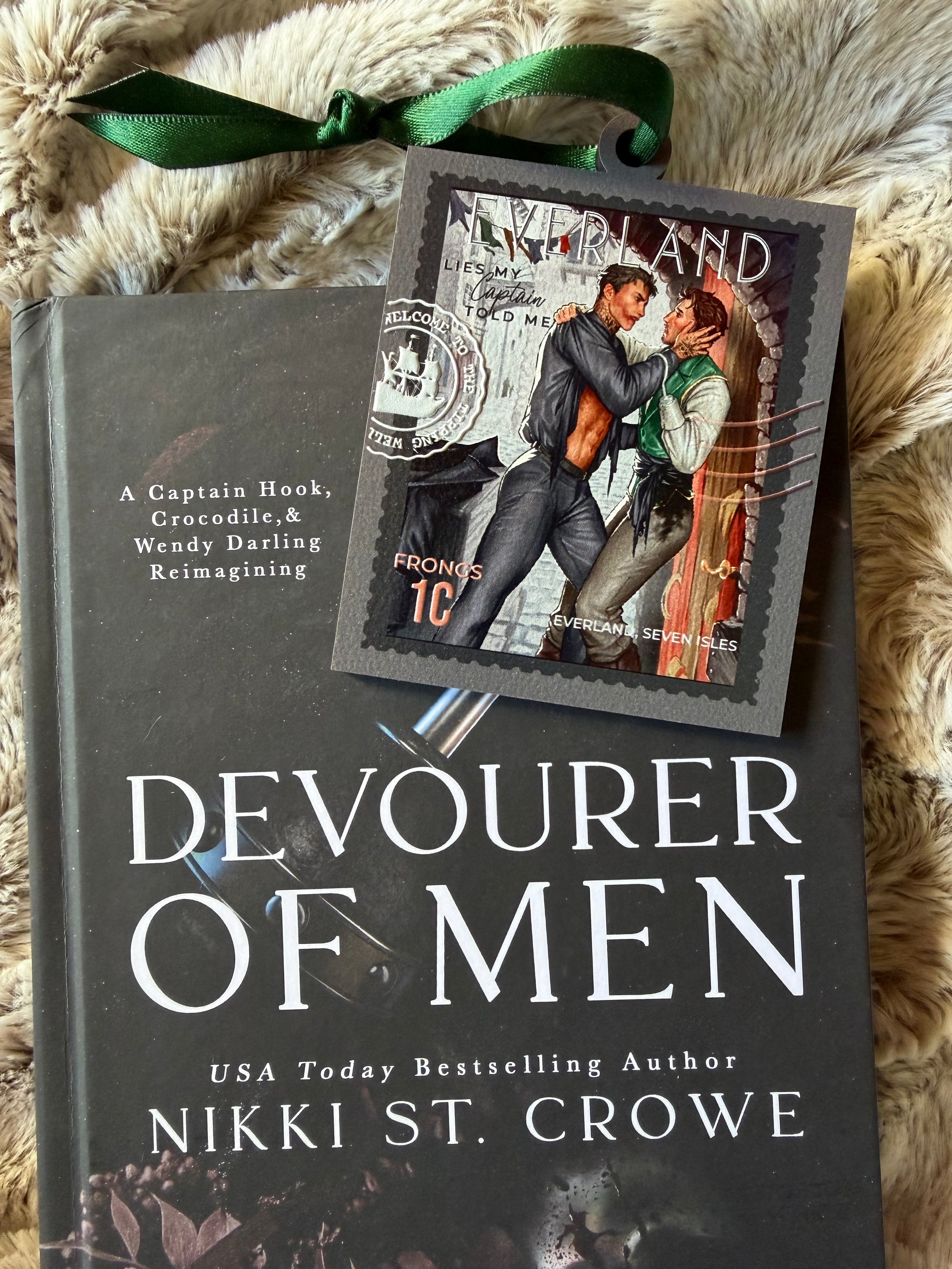 Stamp ornament with "Everland" and a couple in embrace on the top with a green ribbon. Book titled "Devourer of Men" by Nikki St. Crowe in the background. 