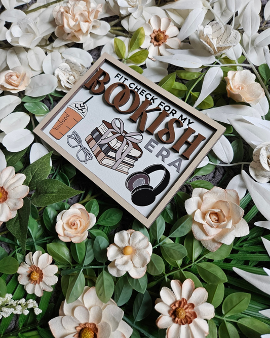 Wooden sign with a white background and tan border. Text reads "Fit Check For My Bookish Era" with flowers in the background. 