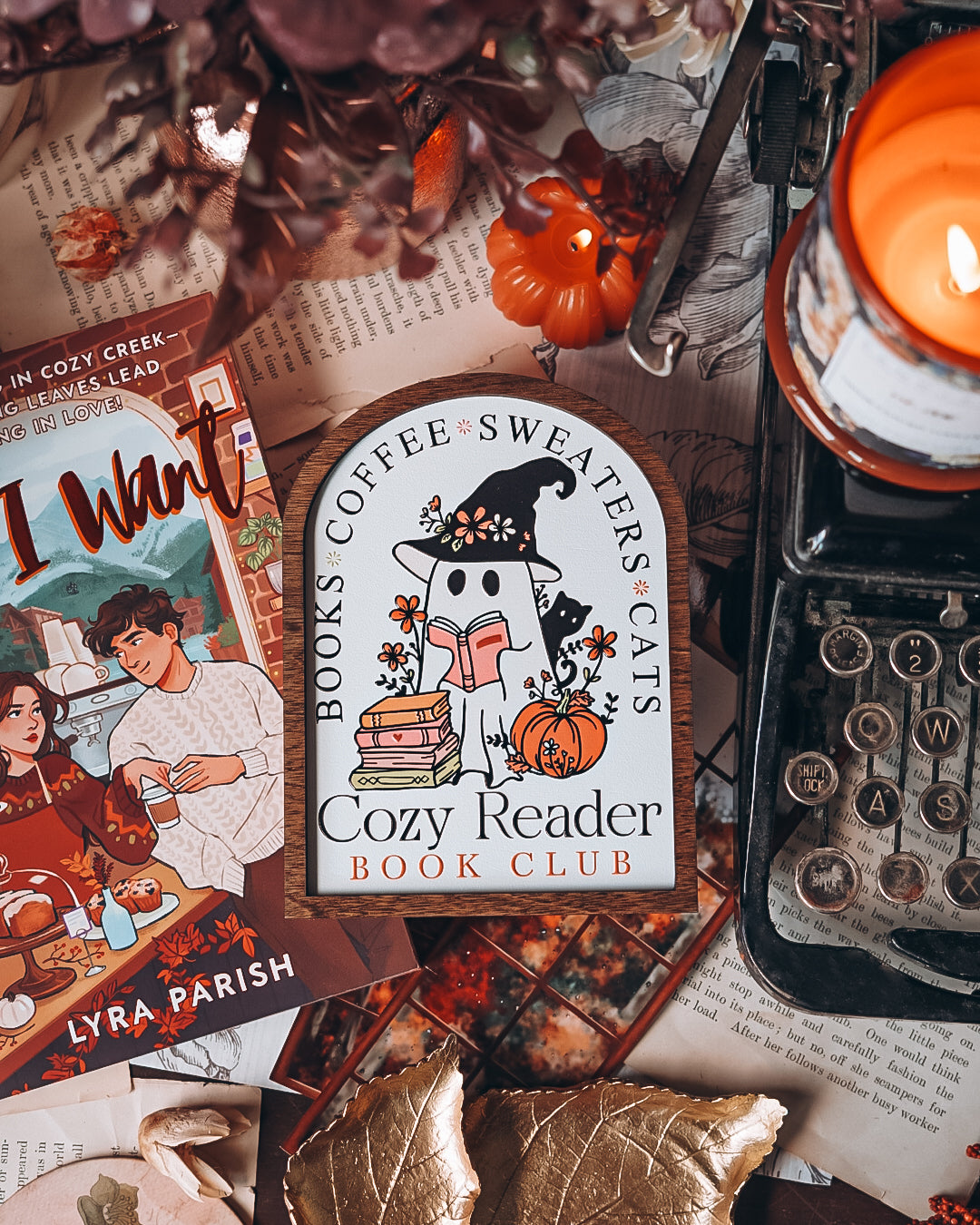 Cozy Reader Ghosty Sign. White background with a wood border with a colorful UV printed design. 
Featuring a ghost reading a book and wearing a witch hat, surrounded by flowers and a pumpkin. Text above reads “Books. Coffee. Sweaters. Cats.” While the text below reads “Cozy Reader Book Club.” 