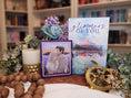 Load image into Gallery viewer, A postage stamp featuring artwork of a romantic scene, in front  of a stack of books, candle, and florals, with library bookshelves in the background. 
