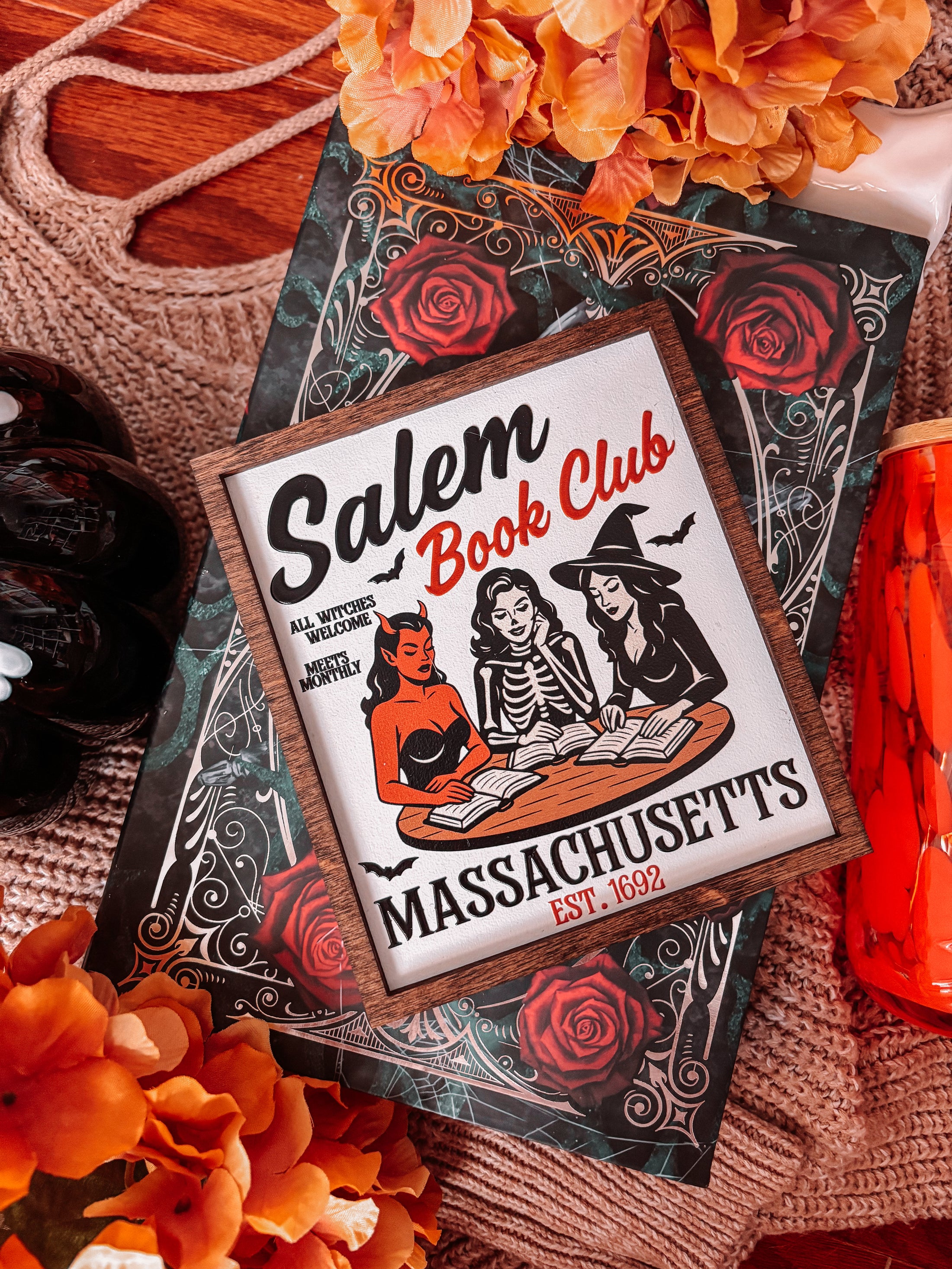 Salem Book Club Sign. White shelf sign with a wood grain border featuring a devil, witch, and skeleton women at a table reading. Top reads “SALEM BOOK CLUB” and the bottom reads “MASSACHUSETTS EST 1692” ©️Fire Drake Artistry 2025